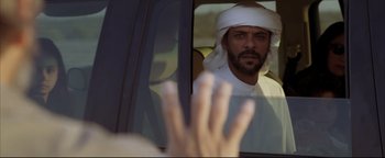 Movie still from “Syriana” (2005), directed by Stephen Gaghan – A man in a white hat is looking in a car mirror; Medium shot, Over the shoulder angle