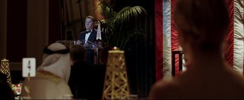 Movie still from “Syriana” (2005), directed by Stephen Gaghan – A man standing at a podium in front of an american flag; Medium shot, Over the shoulder angle