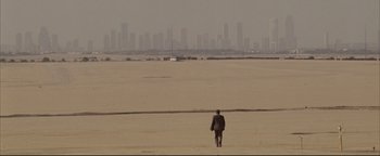 Movie still from “Syriana” (2005), directed by Stephen Gaghan – A man walking across a sandy beach towards a city skyline; Extreme Wide shot, High angle