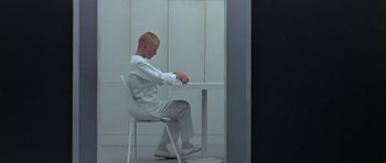 Movie still from “THX 1138” (1971), directed by George Lucas – A man sitting at a table in front of a white wall; Medium shot, High angle