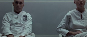 Movie still from “THX 1138” (1971), directed by George Lucas – Two people sitting in a room with a wall behind them; Medium shot, Low angle