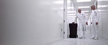 Movie still from “THX 1138” (1971), directed by George Lucas – A person holding a suitcase in a room with white walls; Wide shot, Low angle