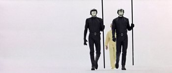 Movie still from “THX 1138” (1971), directed by George Lucas – Two men in black suits and one man in a yellow suit and one man in a black suit; Wide shot, Low angle