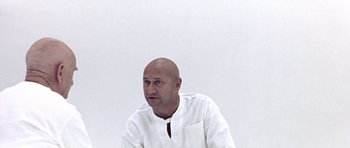 Movie still from “THX 1138” (1971), directed by George Lucas – A bald man in a white shirt is sitting in front of a white wall; Medium shot, Low angle