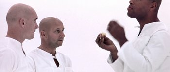 Movie still from “THX 1138” (1971), directed by George Lucas – A man holding a piece of food in front of another man; Close Up shot, Over the shoulder angle