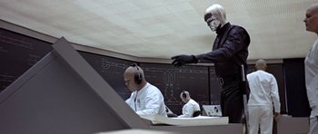Movie still from “THX 1138” (1971), directed by George Lucas – A man in a black suit and a white helmet is standing in front of two other men in a white suit; Medium shot, Low angle