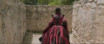 Movie still from “Tale of Tales” (2015), directed by Matteo Garrone – A woman in a red and black dress standing next to a stone wall; Wide shot, Over the shoulder angle