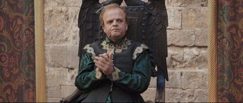 Movie still from “Tale of Tales” (2015), directed by Matteo Garrone – A man with a beard and a chain around his neck is sitting on a chair; Medium shot, High angle