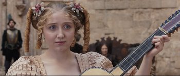 Movie still from “Tale of Tales” (2015), directed by Matteo Garrone – A young woman holding a guitar while standing in front of a stone wall; Close Up shot, Over the shoulder angle