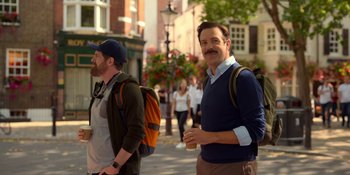 Movie still from “Ted Lasso” (2020), created by Brendan Hunt – A man with a beard and a backpack is standing next to another man; Medium shot, Over the shoulder angle