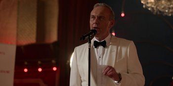 Movie still from “Ted Lasso” (2020), created by Brendan Hunt – An older man in a white suit is speaking into a microphone; Close Up shot, Low angle