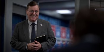 Movie still from “Ted Lasso” (2020), created by Brendan Hunt – A man in a suit and tie holding a book; Medium shot, Over the shoulder angle