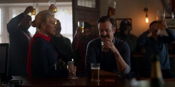 Movie still from “Ted Lasso” (2020), created by Brendan Hunt – A group of people sitting at a table drinking beer; Medium shot, Over the shoulder angle