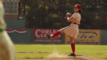Movie still from “A League of Their Own” (2022), created by Abbi Jacobson – A woman in a red and white baseball uniform throwing a ball; Medium shot, Over the shoulder angle