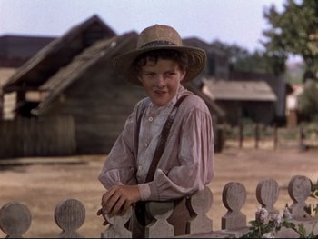 Movie still from “The Adventures of Tom Sawyer” (1938), directed by Norman Taurog – A young boy wearing a straw hat standing next to a wooden fence; Medium shot, Low angle