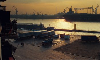 Movie still from “The American Friend” (1977), directed by Wim Wenders – Several trucks parked in a parking lot next to a body of water at sunset; Extreme Wide shot, High angle