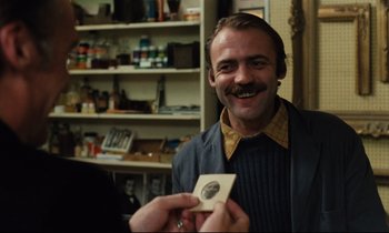 Movie still from “The American Friend” (1977), directed by Wim Wenders – A man with a mustache holding up a piece of paper; Close Up shot, Over the shoulder angle