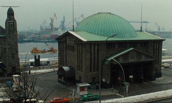 Movie still from “The American Friend” (1977), directed by Wim Wenders – A large building with a green roof in front of a body of water; Extreme Wide shot, High angle