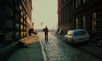 Movie still from “The American Friend” (1977), directed by Wim Wenders – A man walking down a street near a building; Wide shot, High angle