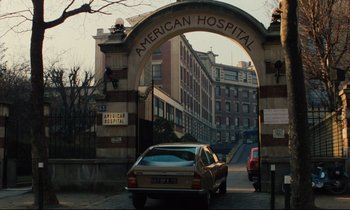 Movie still from “The American Friend” (1977), directed by Wim Wenders – A car driving under an archway in front of an american hospital; Extreme Wide shot, Low angle