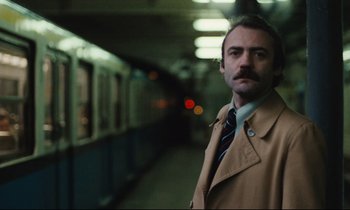 Movie still from “The American Friend” (1977), directed by Wim Wenders – A man in a trench coat standing in front of a train; Close Up shot, Low angle