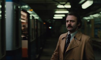 Movie still from “The American Friend” (1977), directed by Wim Wenders – A man in a trench coat standing in a train station; Medium shot, Low angle