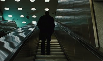 Movie still from “The American Friend” (1977), directed by Wim Wenders – A man is standing on the escalator of a building; Wide shot, Low angle
