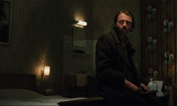 Movie still from “The American Friend” (1977), directed by Wim Wenders – A man sitting in a dark room next to a bed; Medium shot, Low angle