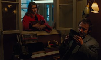 Movie still from “The American Friend” (1977), directed by Wim Wenders – A woman in a red coat looking at a man in a black suit; Medium shot, High angle