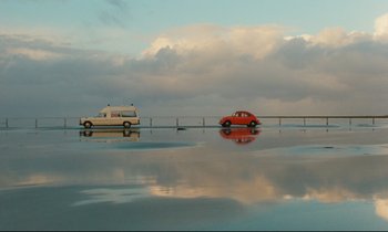Movie still from “The American Friend” (1977), directed by Wim Wenders – A car and a van driving down a road; Extreme Wide shot, Low angle