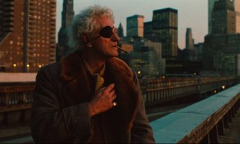 Movie still from “The American Friend” (1977), directed by Wim Wenders – An older man wearing a fur coat and sunglasses with a city in the background; Medium shot, Low angle