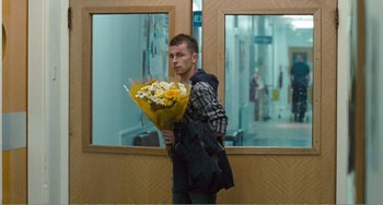 Movie still from “The Angels' Share” (2012), directed by Ken Loach – A man holding a bouquet of flowers in a room; Medium shot, Over the shoulder angle