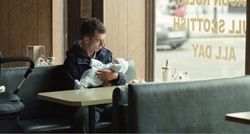 Movie still from “The Angels' Share” (2012), directed by Ken Loach – A man sitting at a table holding a baby; Medium shot, Over the shoulder angle