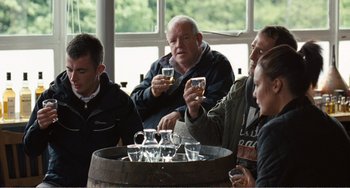 Movie still from “The Angels' Share” (2012), directed by Ken Loach – A group of people sitting around a barrel with glasses of wine; Medium shot, Over the shoulder angle