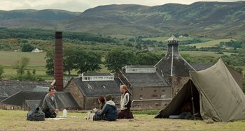 Movie still from “The Angels' Share” (2012), directed by Ken Loach – A group of people sitting on the ground in front of a building; Extreme Wide shot, Over the shoulder angle