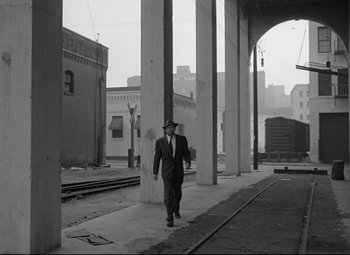 Movie still from “The Asphalt Jungle” (1950), directed by John Huston – A black and white photo of a man in a suit and hat walking on a train platform; Wide shot, Low angle