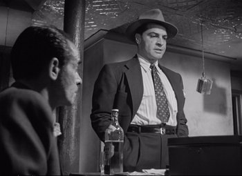 Movie still from “The Asphalt Jungle” (1950), directed by John Huston – A man in a suit and a hat standing in front of a bottle; Medium shot, Low angle