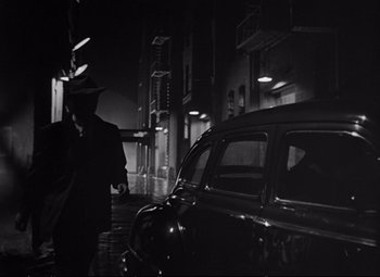 Movie still from “The Asphalt Jungle” (1950), directed by John Huston – A man standing next to a car on a street; Wide shot, Low angle