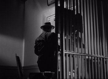 Movie still from “The Asphalt Jungle” (1950), directed by John Huston – A man wearing a hat standing next to a metal gate; Medium shot, Low angle
