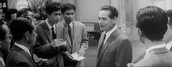 Movie still from “The Bad Sleep Well” (1960), directed by Akira Kurosawa – A man in a suit talks to two other men; Medium shot, Over the shoulder angle