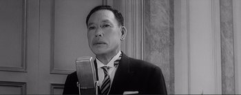 Movie still from “The Bad Sleep Well” (1960), directed by Akira Kurosawa – A man in a suit and tie holding a microphone; Close Up shot, Low angle