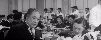 Movie still from “The Bad Sleep Well” (1960), directed by Akira Kurosawa – A group of people sitting at a table with food; Medium shot, Over the shoulder angle