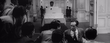 Movie still from “The Bad Sleep Well” (1960), directed by Akira Kurosawa – A group of people standing in a room with a large cake; Medium shot, Over the shoulder angle