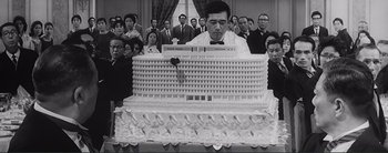 Movie still from “The Bad Sleep Well” (1960), directed by Akira Kurosawa – A man in front of a large cake in front of a group of people; Medium shot, Low angle