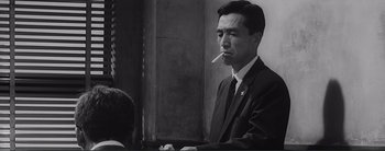 Movie still from “The Bad Sleep Well” (1960), directed by Akira Kurosawa – A man smoking a cigarette while sitting at a table in front of other people; Close Up shot, Low angle