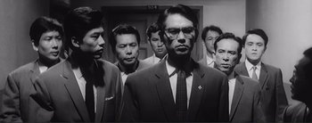Movie still from “The Bad Sleep Well” (1960), directed by Akira Kurosawa – A group of men standing next to each other in front of a door; Medium shot, Low angle