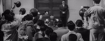 Movie still from “The Bad Sleep Well” (1960), directed by Akira Kurosawa – A man in a suit and tie sitting in front of a crowd; Medium shot, Over the shoulder angle