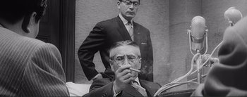 Movie still from “The Bad Sleep Well” (1960), directed by Akira Kurosawa – An older man smoking a cigarette in front of an older man; Medium shot, Low angle