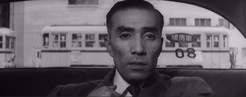 Movie still from “The Bad Sleep Well” (1960), directed by Akira Kurosawa – A man in a suit and tie in front of a building; Close Up shot, Low angle