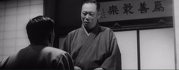 Movie still from “The Bad Sleep Well” (1960), directed by Akira Kurosawa – An older man wearing a robe in front of an oriental sign; Medium shot, Over the shoulder angle
